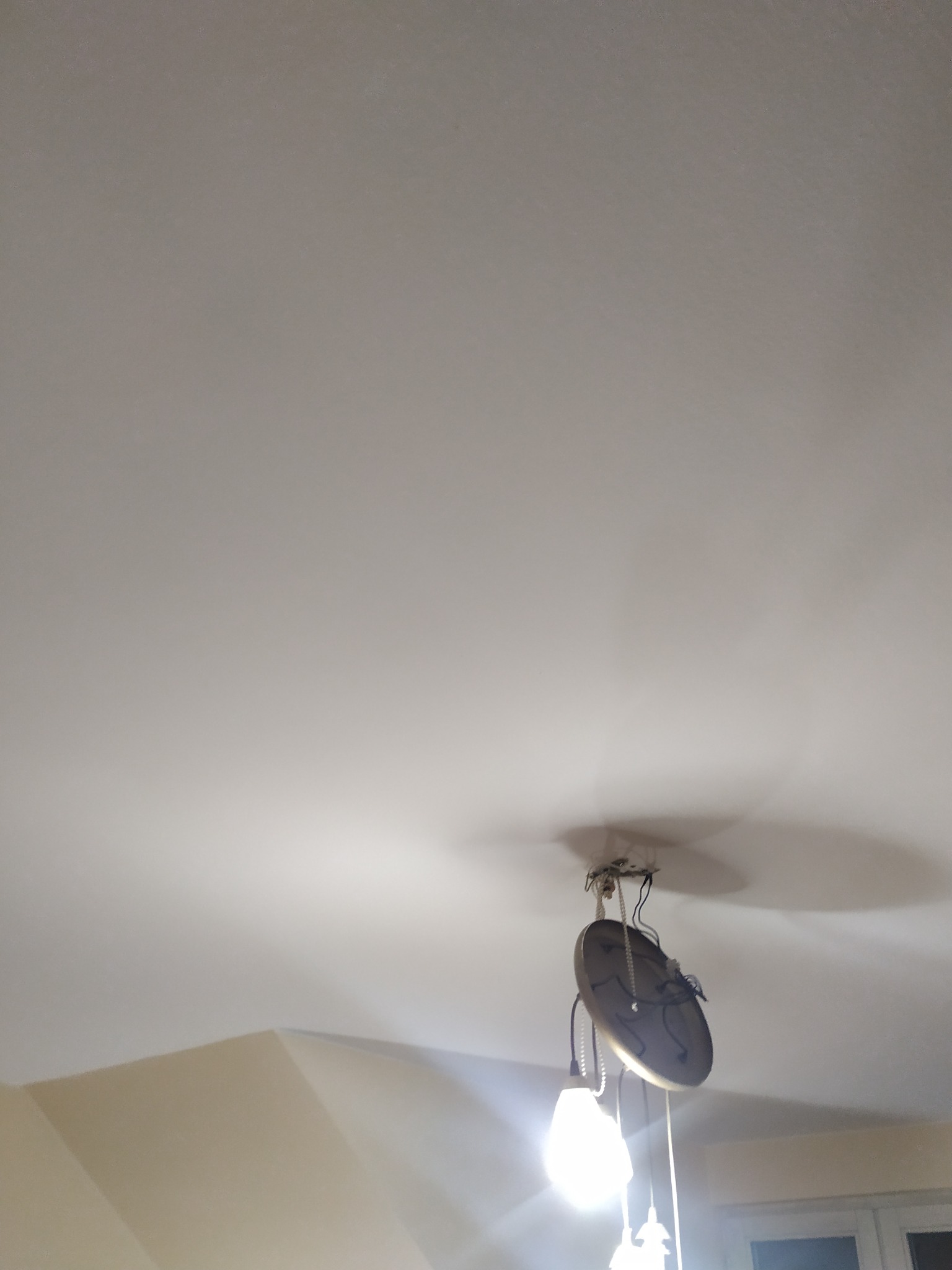 Biały sufit po malowaniu z widoczną lampą wiszącą. Świeżo pomalowany sufit w jasnym odcieniu z widocznymi detalami instalacji oświetleniowej.