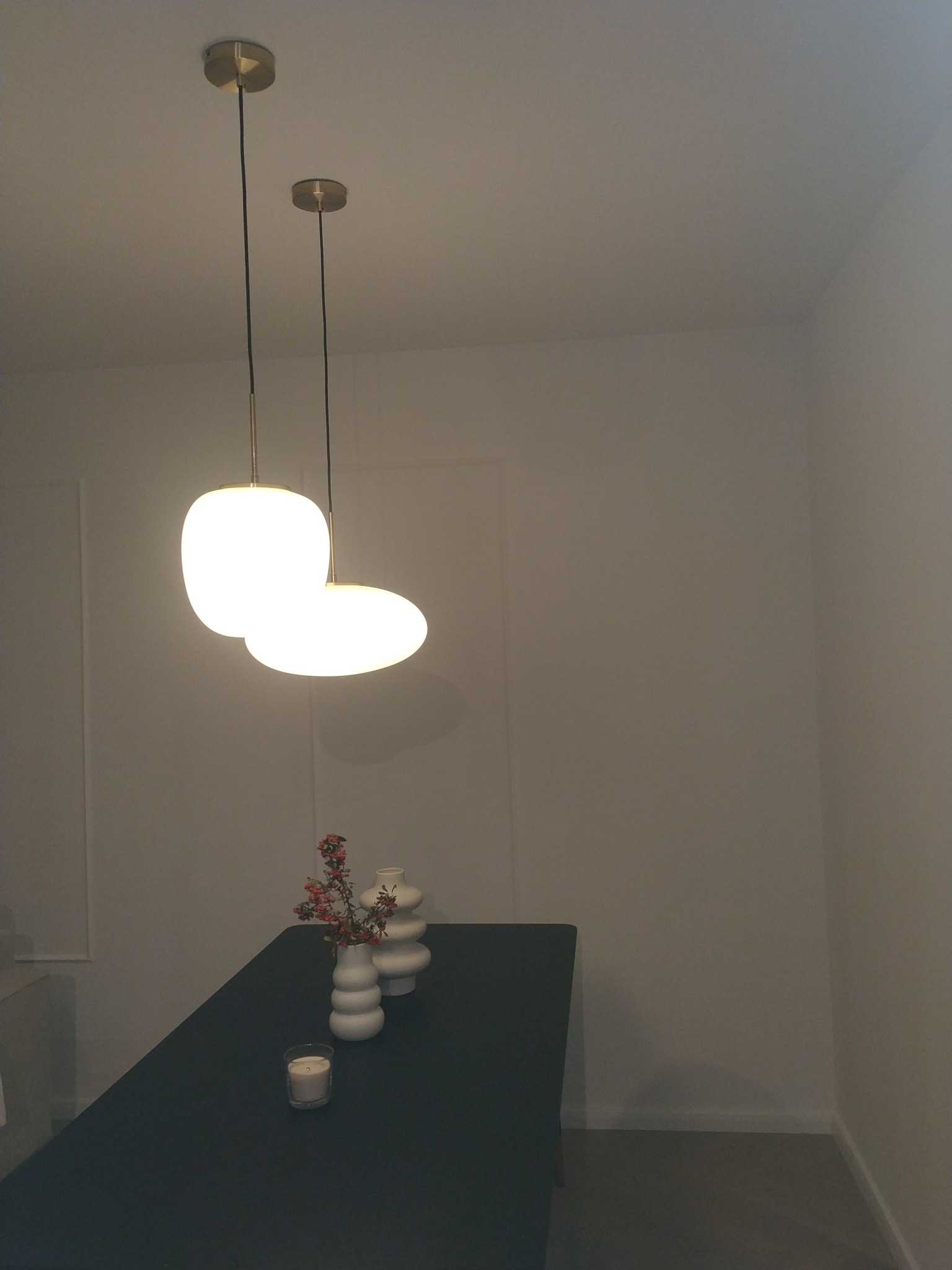 Lampa wisząca z dwoma białymi kloszami nad ciemnym stołem, na którym stoją białe wazony z kwiatami i zapalona świeczka. Prosty, minimalistyczny design wnętrza.