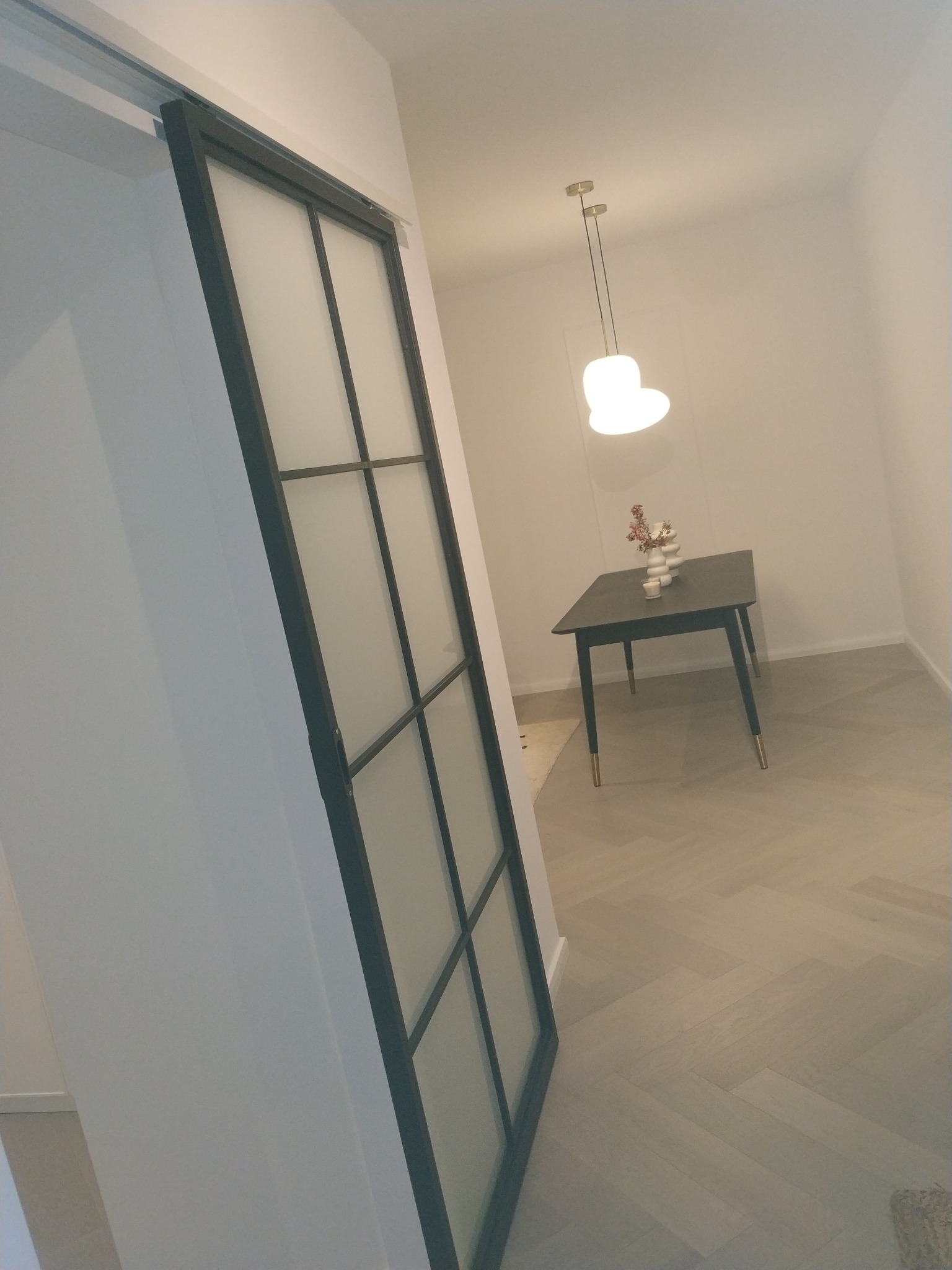 Nowoczesne wnętrze z przesuwnymi drzwiami szklanymi z czarną ramą, widok na stół z dekoracjami i lampą wiszącą. Parkiet w jodełkę.