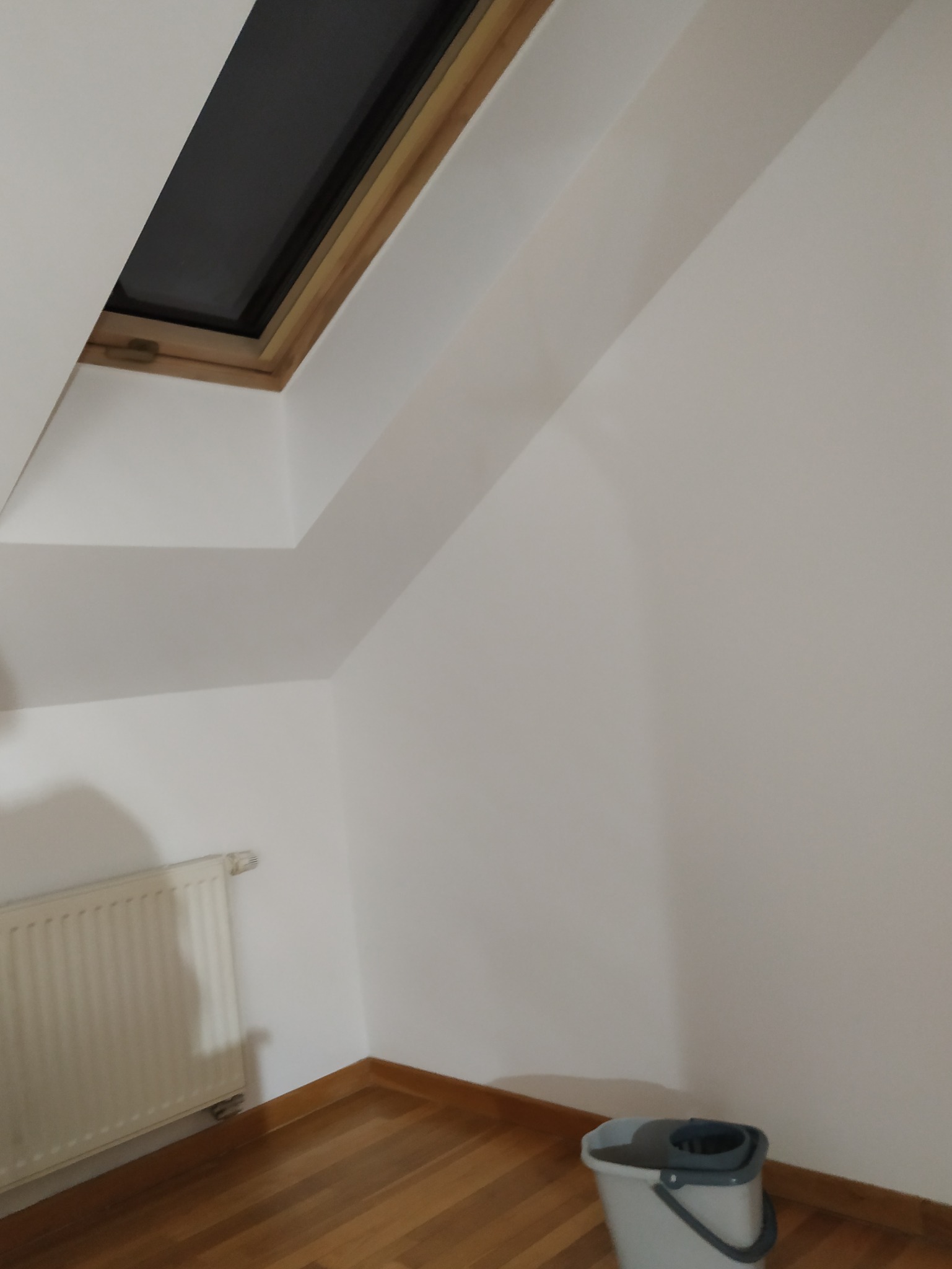 Białe ściany poddasza z oknem dachowym, grzejnikiem i mopem. Świeżo pomalowane, gładkie powierzchnie. Parkiet na podłodze. Widok z dołu.