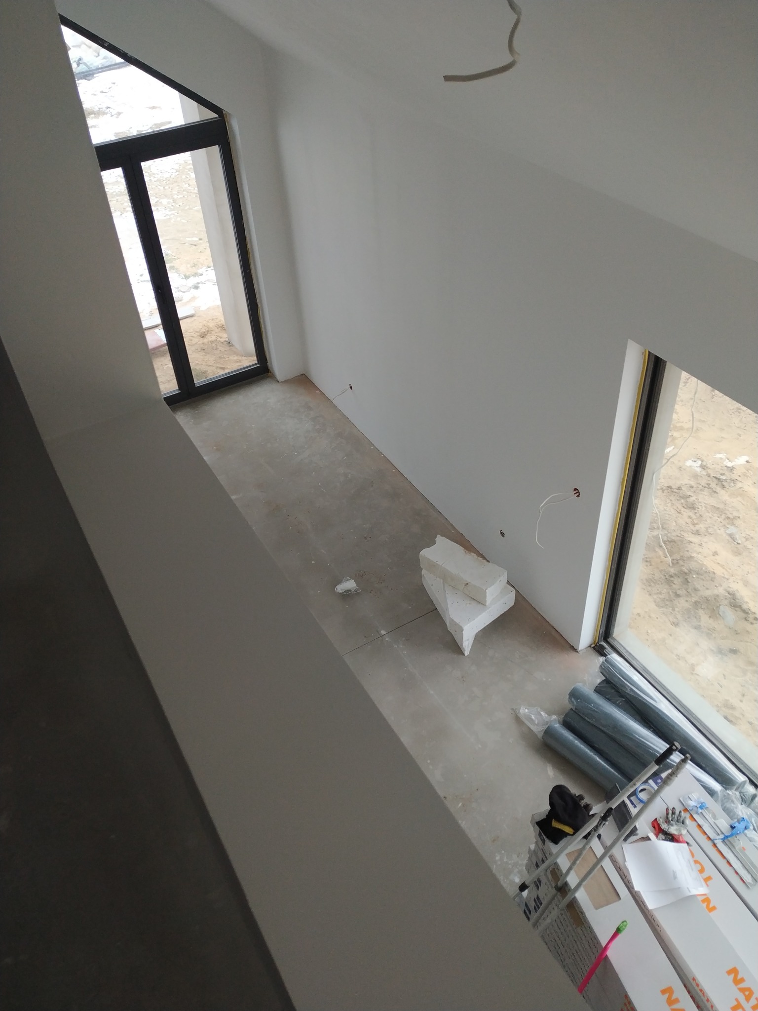 Wnętrze w trakcie prac wykończeniowych: białe ściany, okna z widokiem na zewnątrz, materiały budowlane i drabina. Widok z góry, surowy stan przed malowaniem.