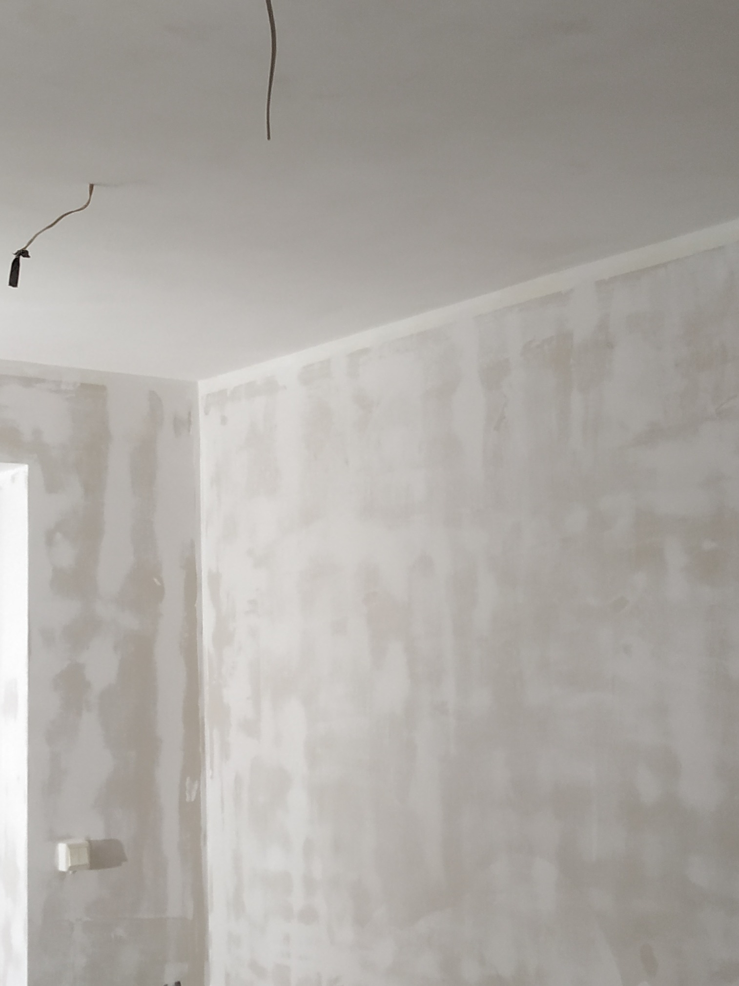 Ściana po nałożeniu gładzi, przygotowana do malowania. Widoczne przewody elektryczne wystające z sufitu. Ściany w jasnych, neutralnych odcieniach szarości i bieli.