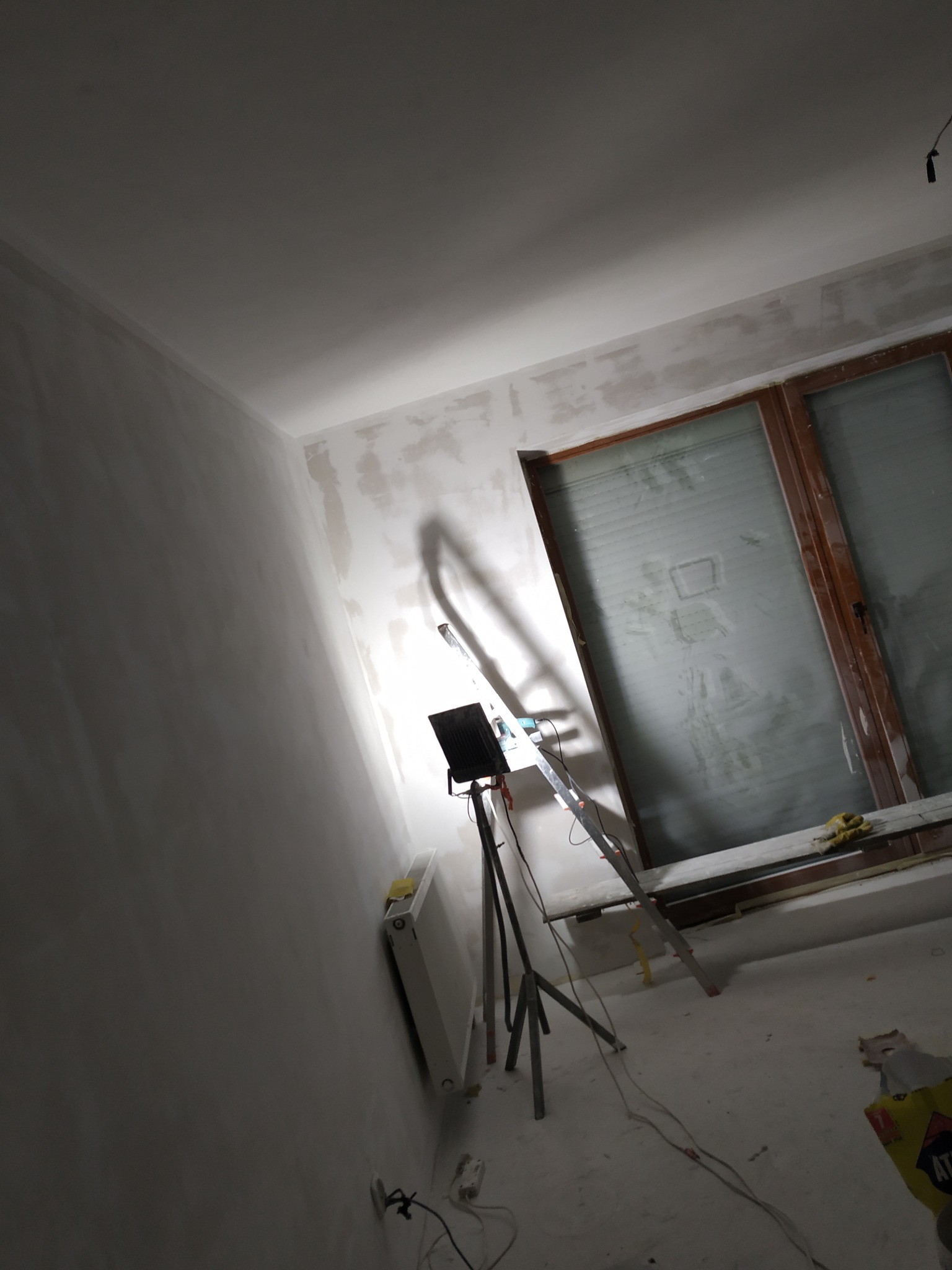 Ściana po nałożeniu gładzi, przygotowana do malowania. Widoczna drabina, lampa oświetlająca ścianę i okno zabezpieczone folią. Ukośne ujęcie.