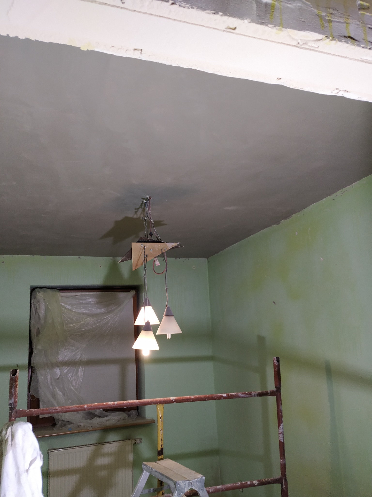 Pokój w trakcie remontu: świeżo położona gładź na suficie, ściany w odcieniach zieleni, okno zabezpieczone folią, widoczne rusztowanie i wisząca lampa.