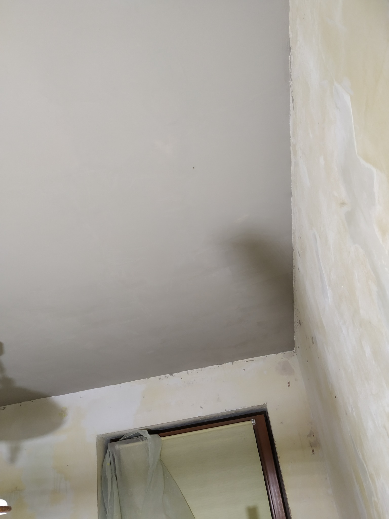 Świeżo położona, jasnoszara gładź na suficie i ścianie w rogu pokoju, widoczne okno z zasłoniętą szybą. Fragment ściany przed gładzią. Ujęcie z dołu, podkreślające gładką powierzchnię.