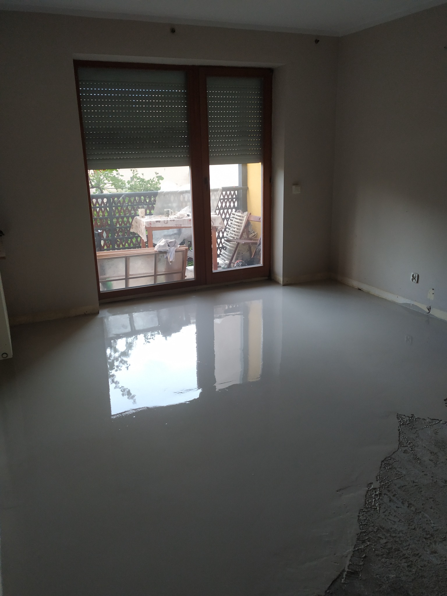 Wylana, mokra posadzka samopoziomująca w pokoju z oknem balkonowym. Widoczne odbicie okna w mokrej powierzchni. Część podłogi jeszcze niezalana.