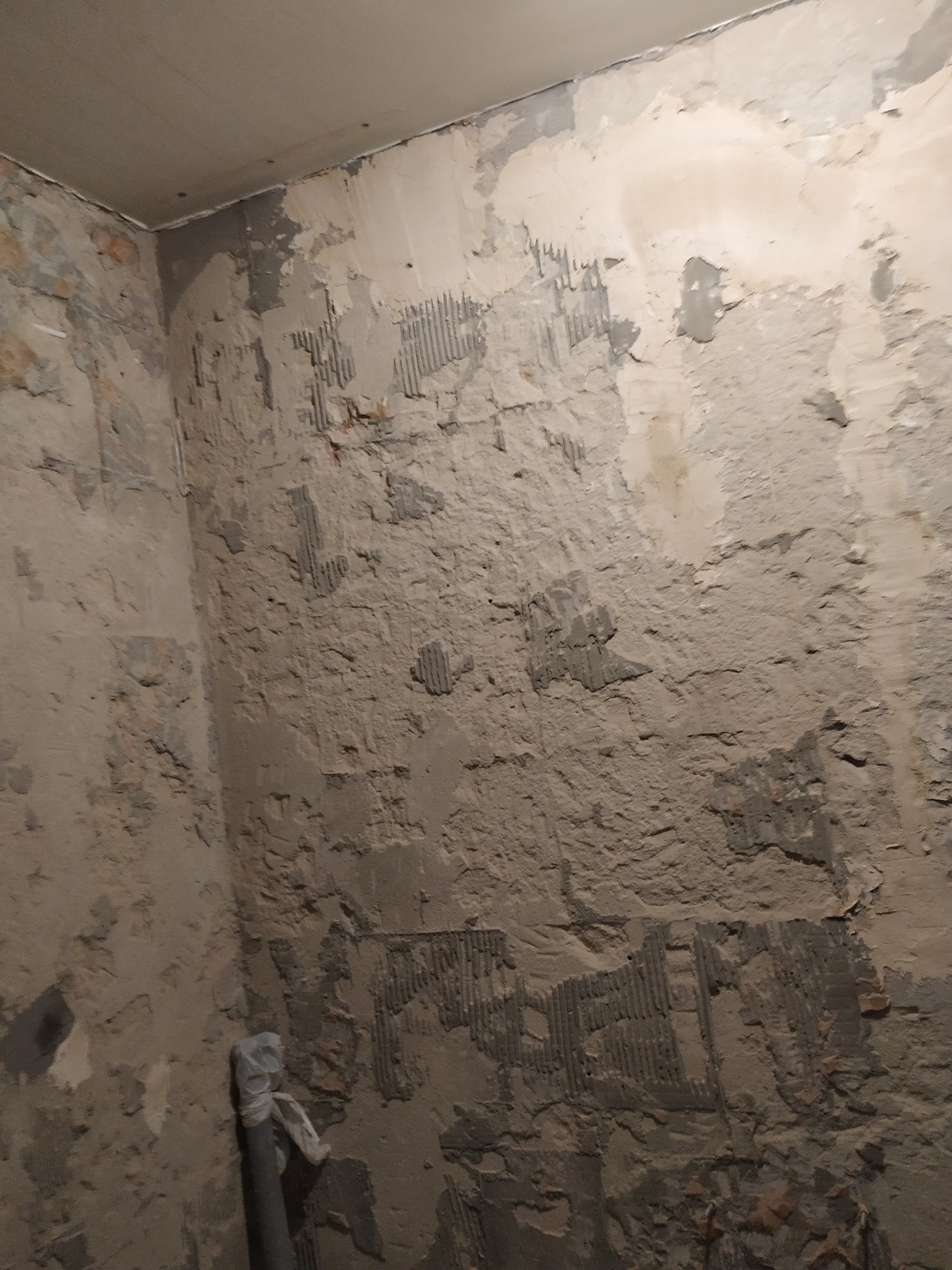 Surowe ściany w trakcie remontu, widoczne ślady po płytkach i nierówności. Przygotowanie pod gładzie lub malowanie, rura w rogu pomieszczenia.