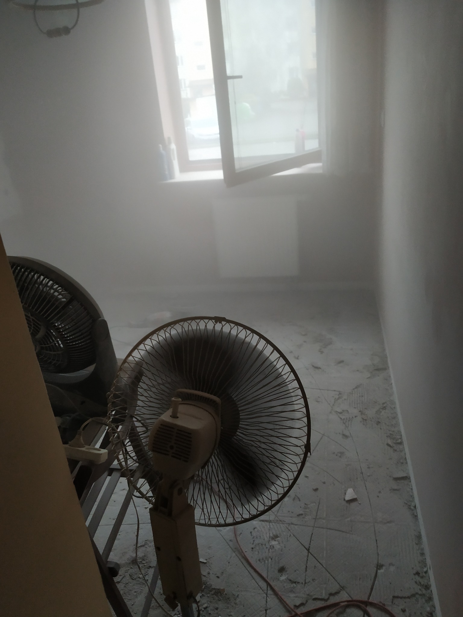 Pokój w trakcie remontu, widoczne dwa wentylatory stojące na podłodze, zapylone powietrze, ślady po gładzi na ścianach i podłodze, okno w tle.