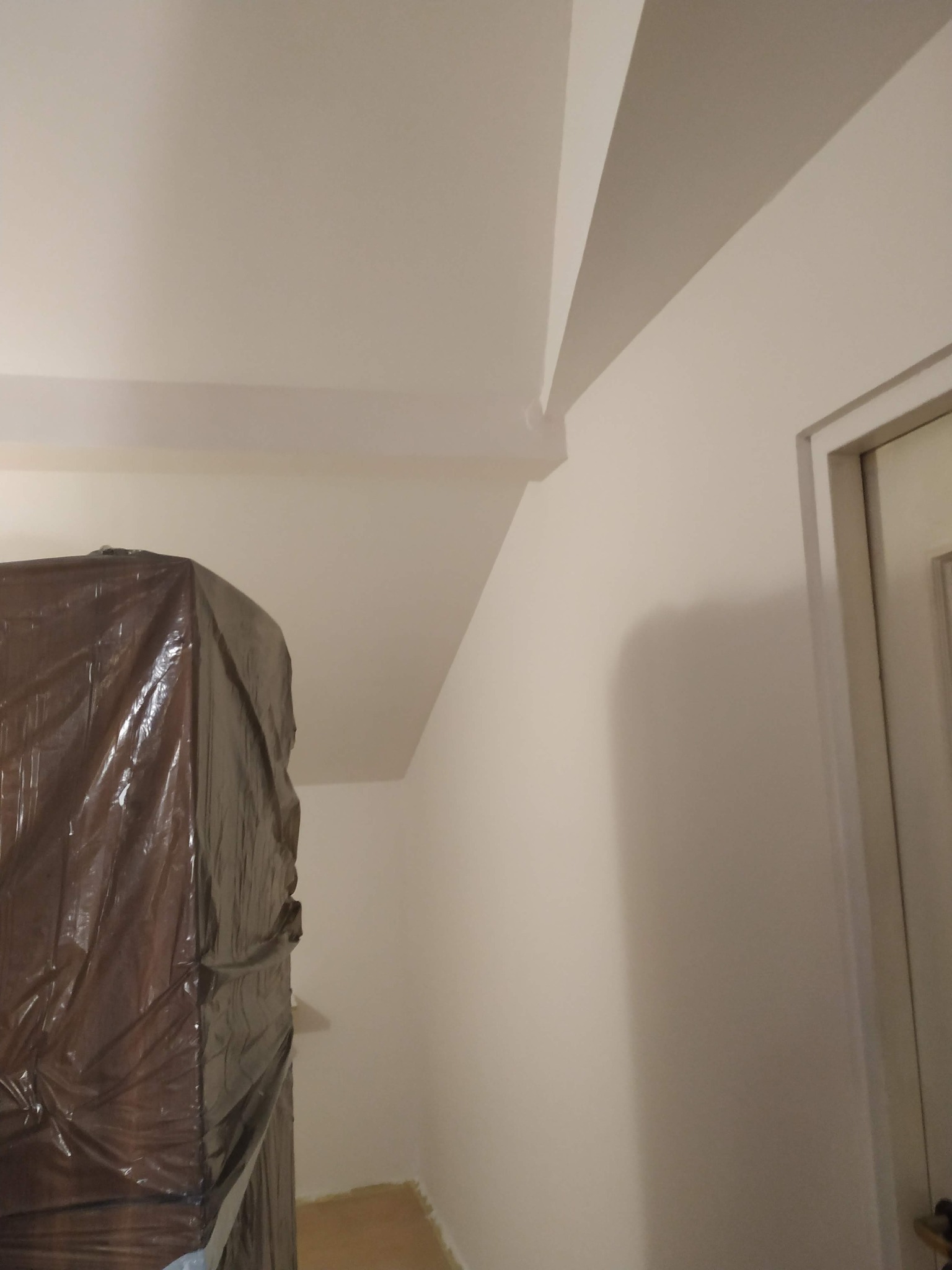 Pomalowane na biało ściany i sufit w pokoju, zabezpieczone meble folią malarską, widoczny fragment drzwi. Świeżo po malowaniu.
