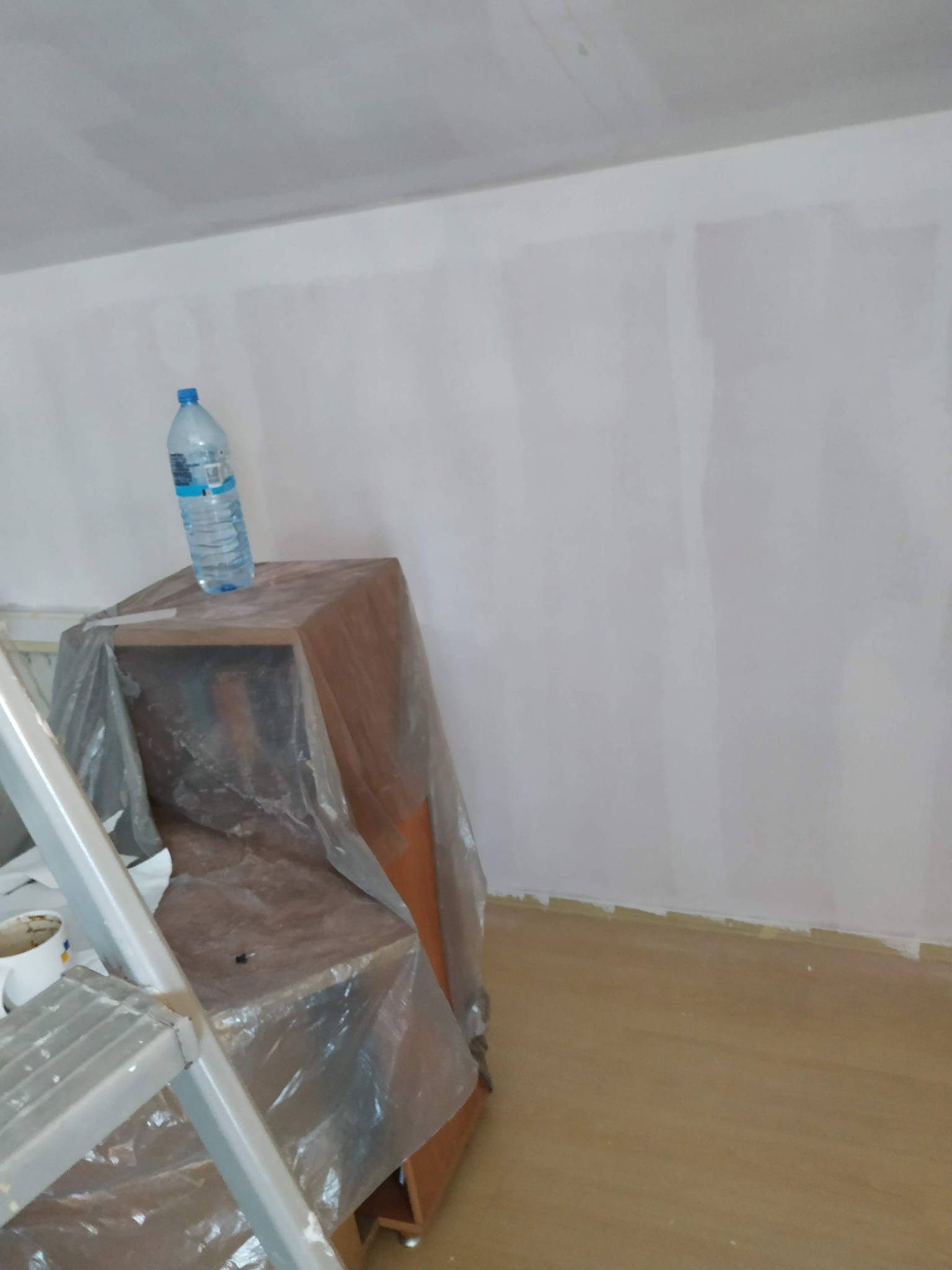 Świeżo pomalowana na biało ściana w pokoju, zabezpieczone meble folią, drabina i butelka wody. Widoczne niedoskonałości w malowaniu przy podłodze.