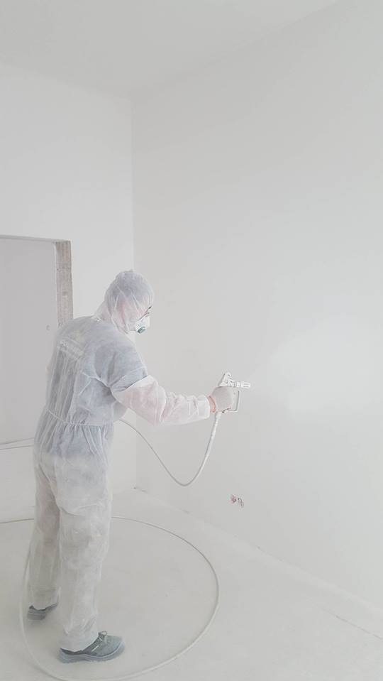 Pracownik w kombinezonie ochronnym maluje ścianę na biało za pomocą pistoletu natryskowego. Widoczne gniazdka elektryczne na ścianie.