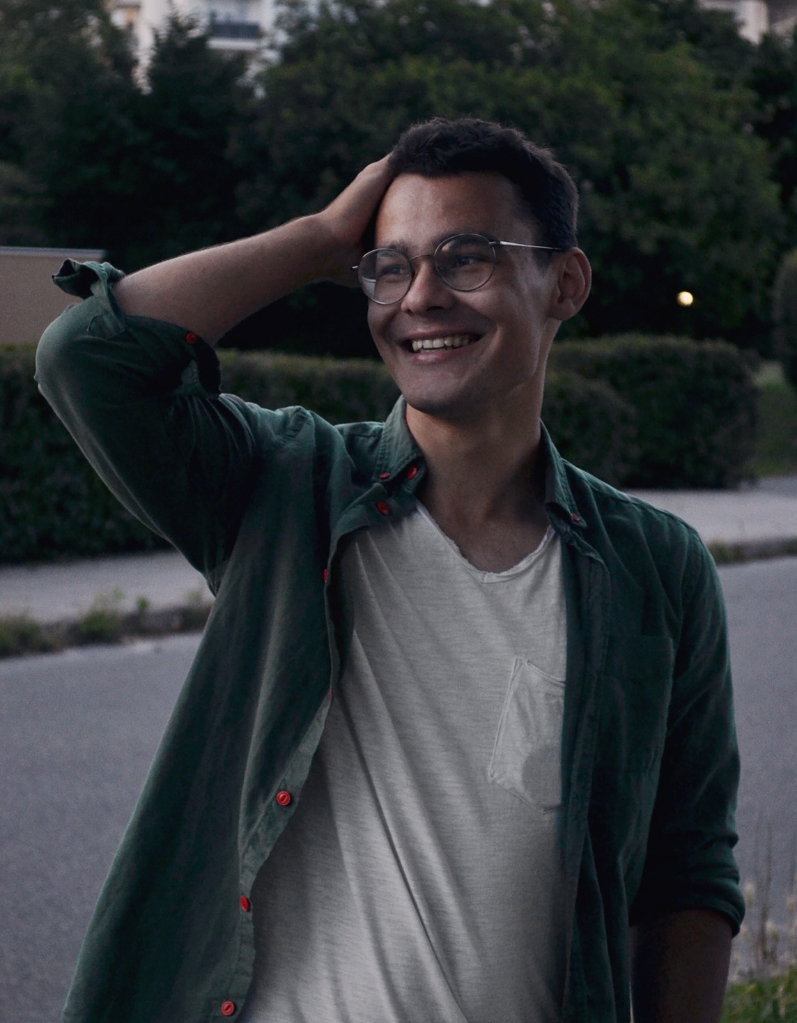 Uśmiechnięty mężczyzna w okularach i zielonej koszuli dotyka włosów na tle zieleni miejskiej i asfaltowej drogi. Widok z dołu, naturalne światło, ekspresja radości.