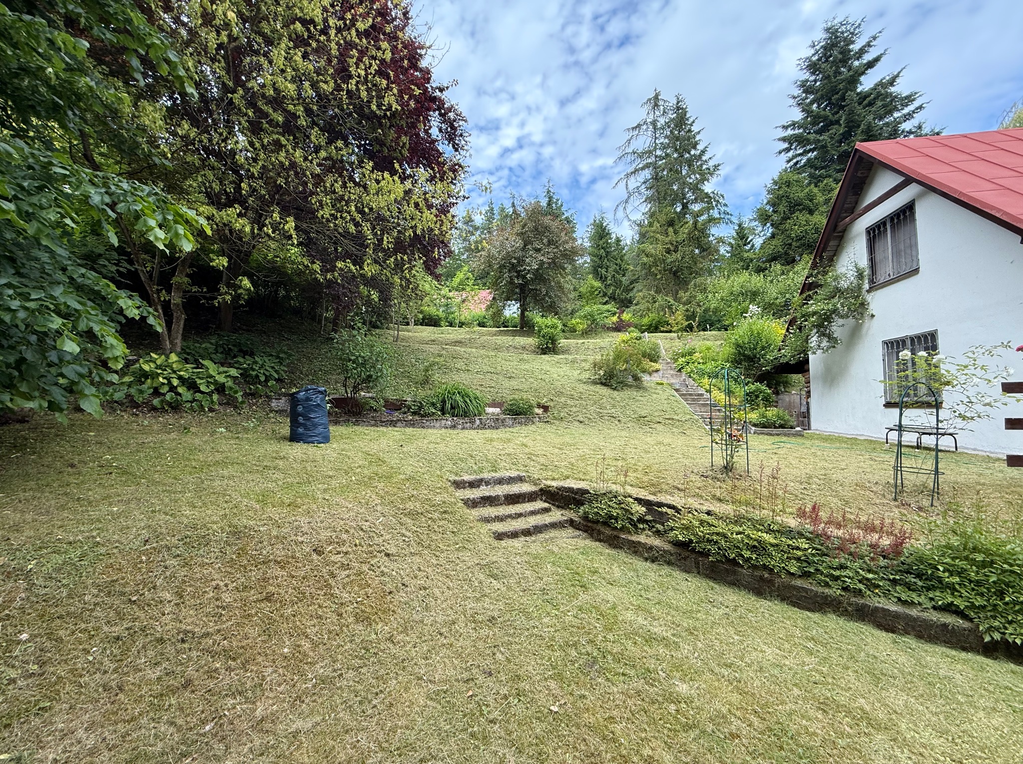Działka po koszeniu trawy: równy trawnik z wzniesieniami, rabatami i schodami, widoczny worek z trawą. W tle dom z czerwoną dachówką i drzewa.