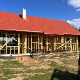 Dom w stanie surowym otwartym z czerwoną dachówką i białą konstrukcją komina na tle błękitnego nieba. Widoczna drewniana więźba dachowa i rynny.