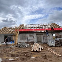 PabloEnergy - Budowa domu w toku: widoczna więźba dachowa z czerwoną membraną, pracownicy na dachu, teren budowy z błotem i materiałami budowlanymi.