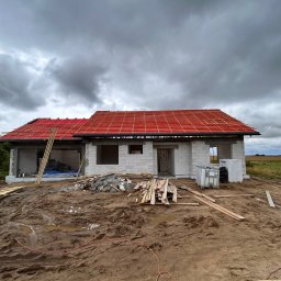 PabloEnergy - Dom w trakcie budowy, z częściowo ułożoną czerwoną dachówką na łatach. Widoczne materiały budowlane i błotniste podłoże. Pochmurne niebo w tle.