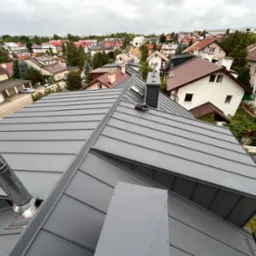 Szary, blaszany dach z kominami na tle zabudowy miejskiej w pochmurny dzień. Widok z g&oacute;ry na nową konstrukcję.