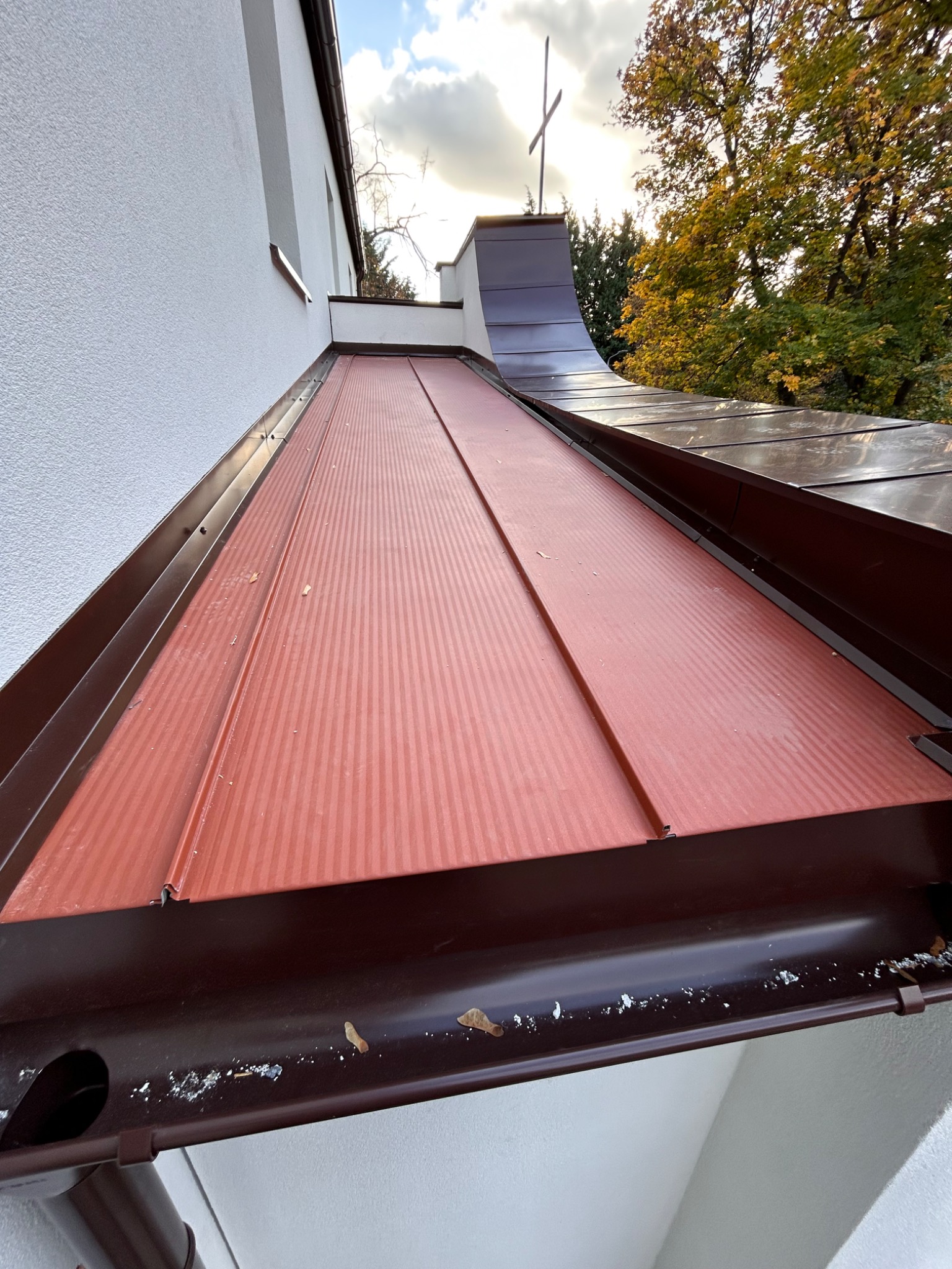 Czerwono-brązowe, blaszane pokrycie dachu z widocznymi elementami orynnowania, na tle fragmentu białej ściany budynku i jesiennych drzew.
