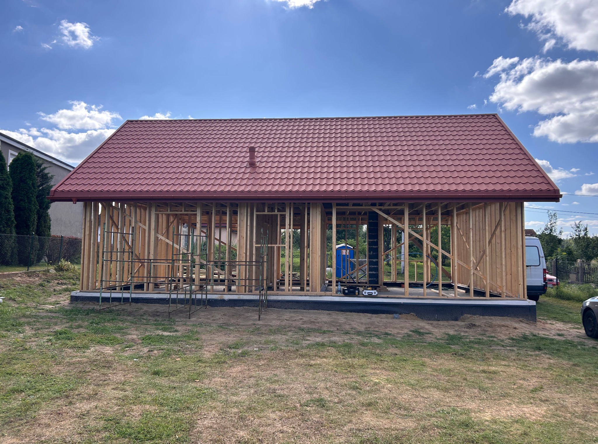 Budowa domu w trakcie: drewniana konstrukcja z czerwonym dachem na tle błękitnego nieba. Widoczne rusztowania i narzędzia.