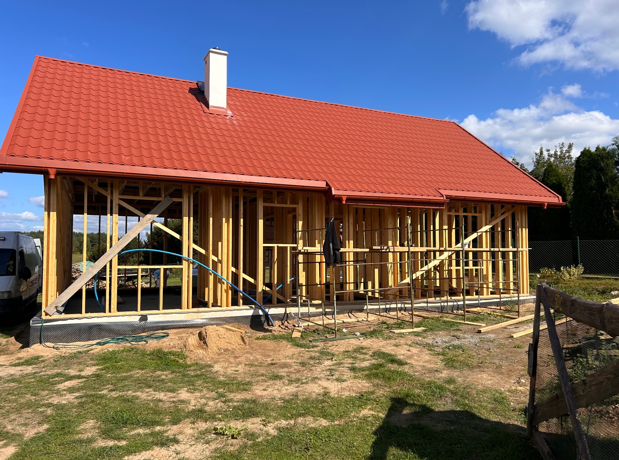 Dom w stanie surowym otwartym z czerwoną dachówką i białą konstrukcją komina na tle błękitnego nieba. Widoczna drewniana więźba dachowa i rynny.