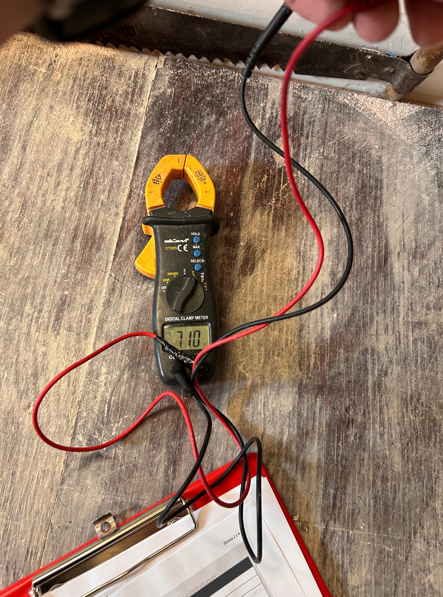 Miernik cęgowy z przewodami na drewnianej powierzchni, wskazujący 710 V, obok dokument w czerwonej oprawie. Pomiar napięcia w instalacji elektrycznej.