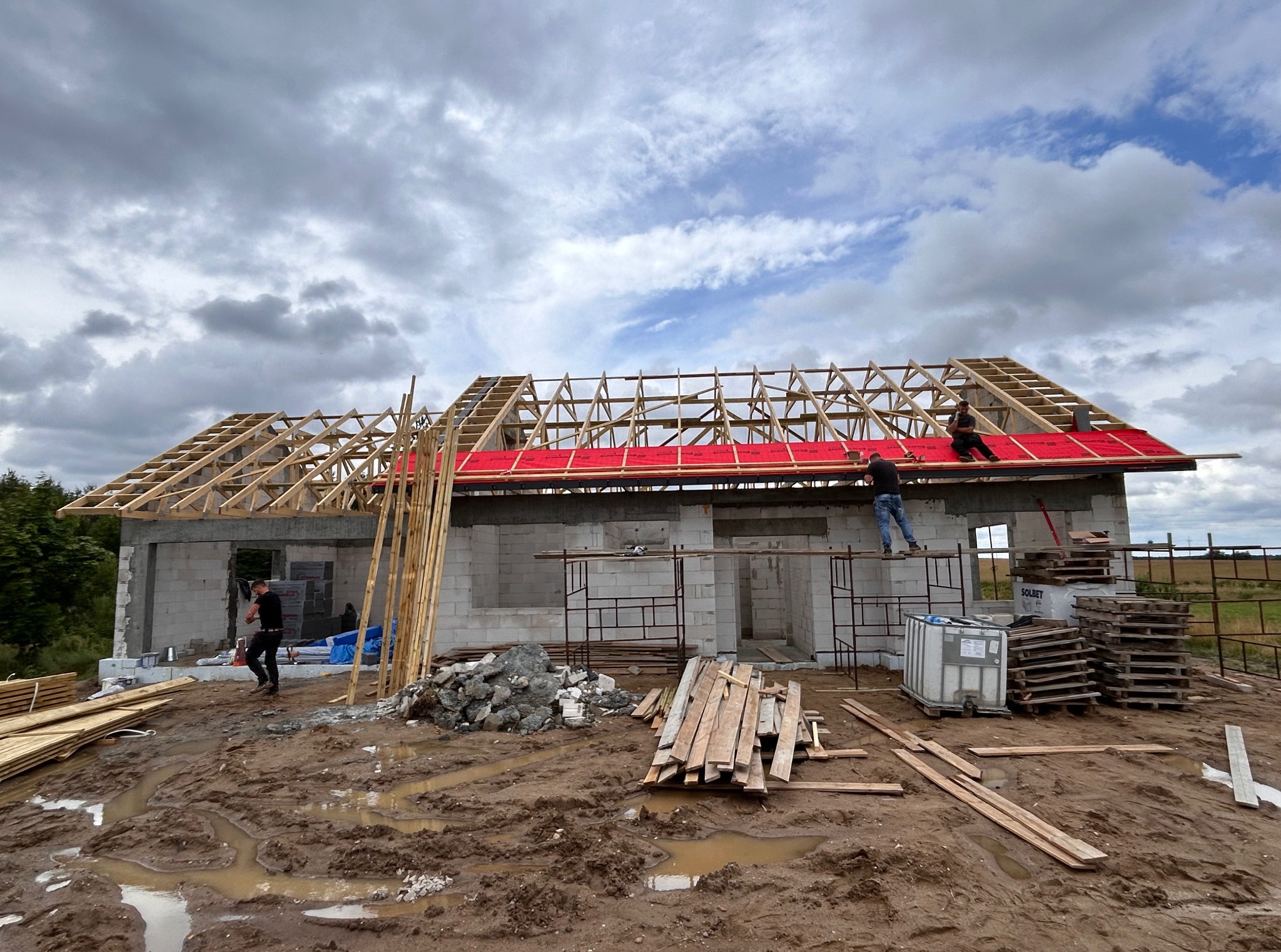 Budowa domu w toku: widoczna więźba dachowa z czerwoną membraną, pracownicy na dachu, teren budowy z błotem i materiałami budowlanymi.