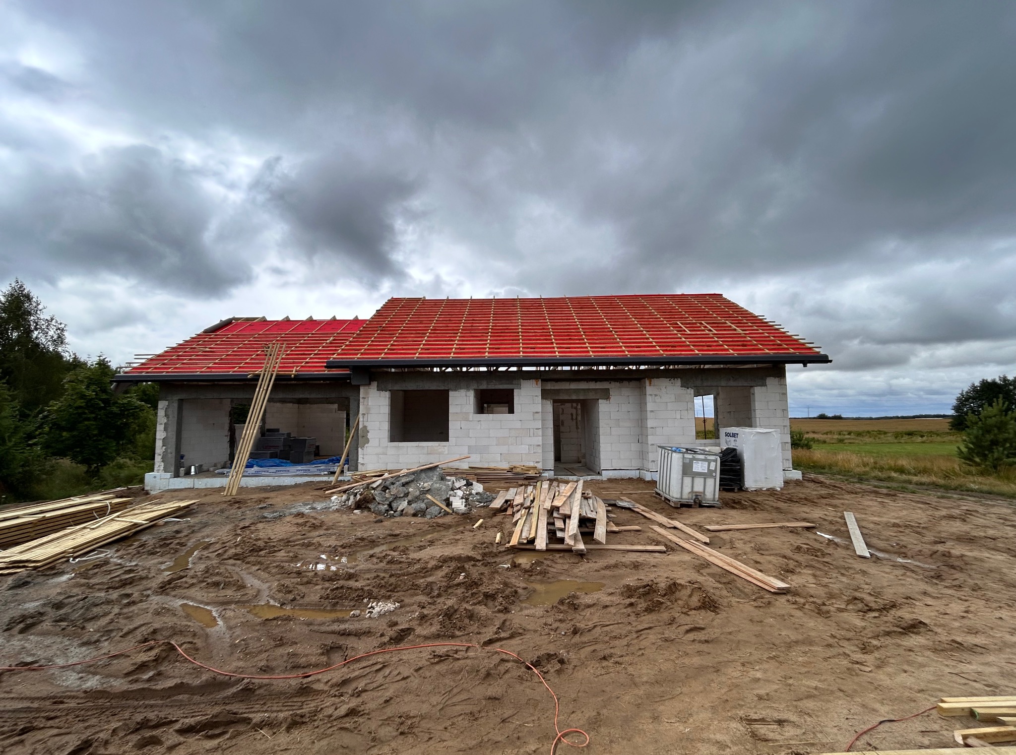 Dom w trakcie budowy, z częściowo ułożoną czerwoną dachówką na łatach. Widoczne materiały budowlane i błotniste podłoże. Pochmurne niebo w tle.