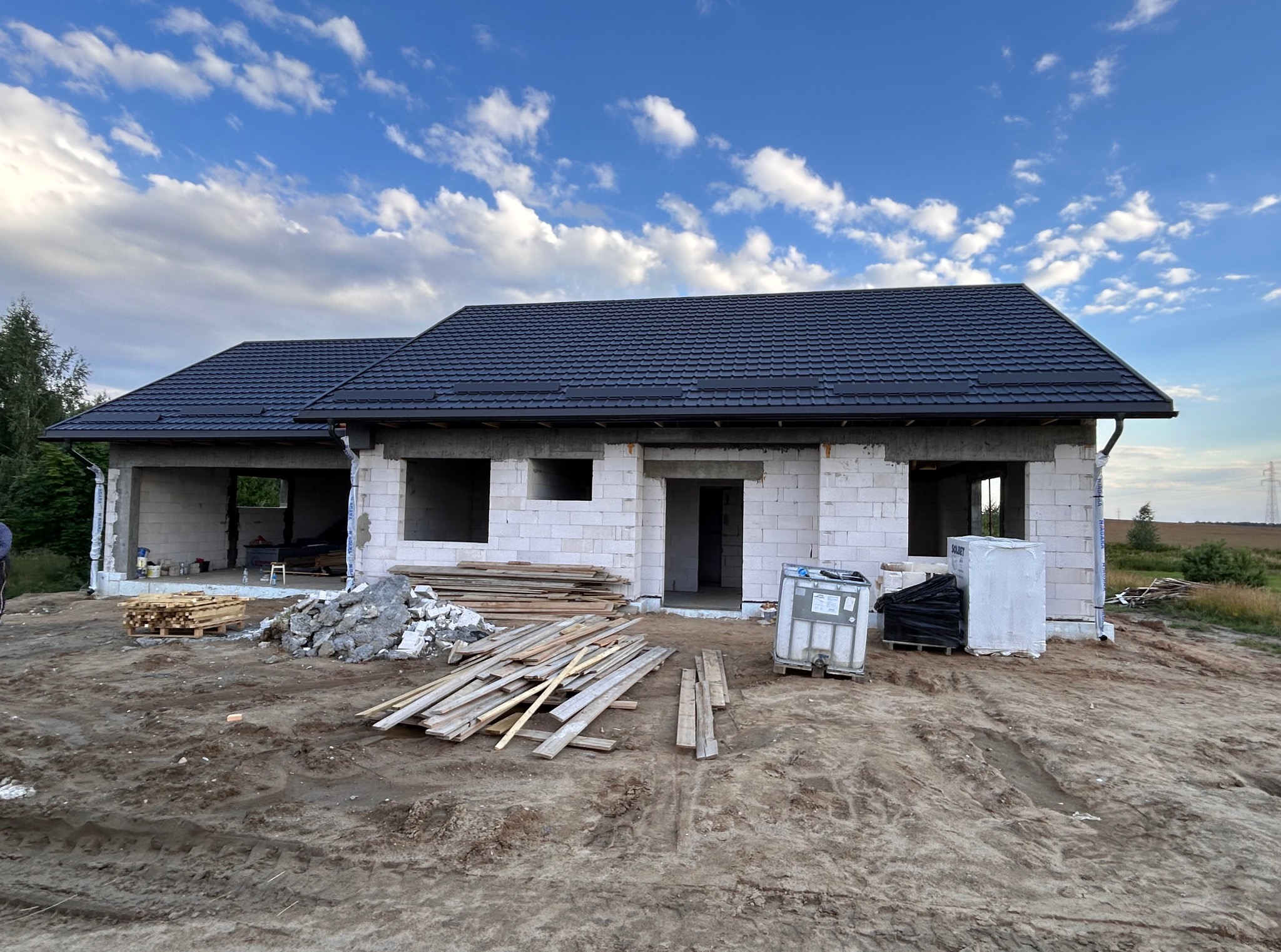 Dom w trakcie budowy z ciemnym dachem na tle błękitnego nieba z chmurami. Widoczne materiały budowlane i surowe ściany z pustaków.