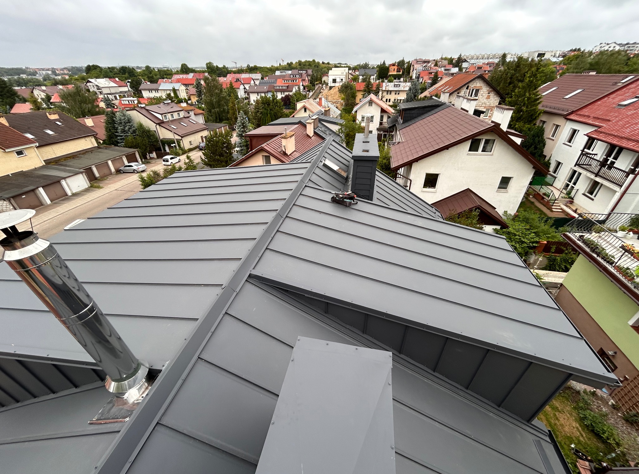 Szary, blaszany dach z kominami na tle zabudowy miejskiej w pochmurny dzień. Widok z góry na nową konstrukcję.