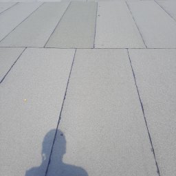 Waterproofing - Szare, płaskie płyty tworzące dach, z widoczną cieniowaną sylwetką osoby w dolnej części kadru i narzędziami dekarskimi w tle, ukazujące etap prac.