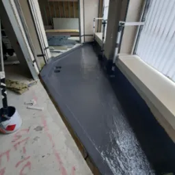 Hydroizolacja balkonu w trakcie prac: świeżo nałożona, ciemnoszara warstwa izolacyjna na podłożu, widoczne okno i ściany w surowym stanie. Wiadro z farbą w rogu.