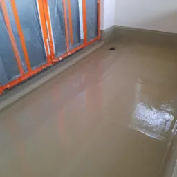 Wnętrze po wylewce samopoziomującej, z zabezpieczonymi folią i taśmą oknami balkonowymi. Widoczny odpływ liniowy i świeżo wylana, gładka powierzchnia.