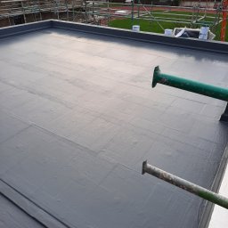 Waterproofing - Szary, płaski dach po renowacji z widocznymi śladami wałka, z zielonymi podpórkami rusztowania i widokiem na morze w tle. Widoczne puszki z farbą.