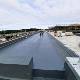 Waterproofing - Szary, płaski dach po hydroizolacji z widocznymi kominami wentylacyjnymi i rusztowaniami w tle. Świeżo położona powłoka, brak detali wykończeniowych.