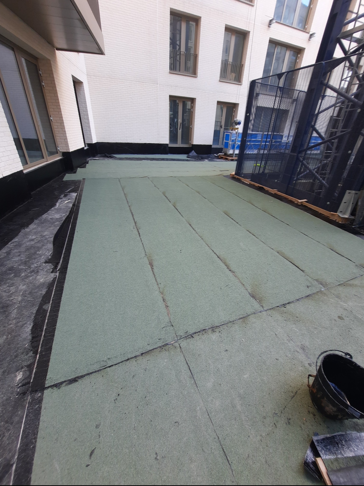 Płaski dach pokryty zielonymi panelami izolacyjnymi, widoczne narzędzia dekarskie i fragment rusztowania. Biały budynek w tle. Etap prac izolacyjnych dachu.