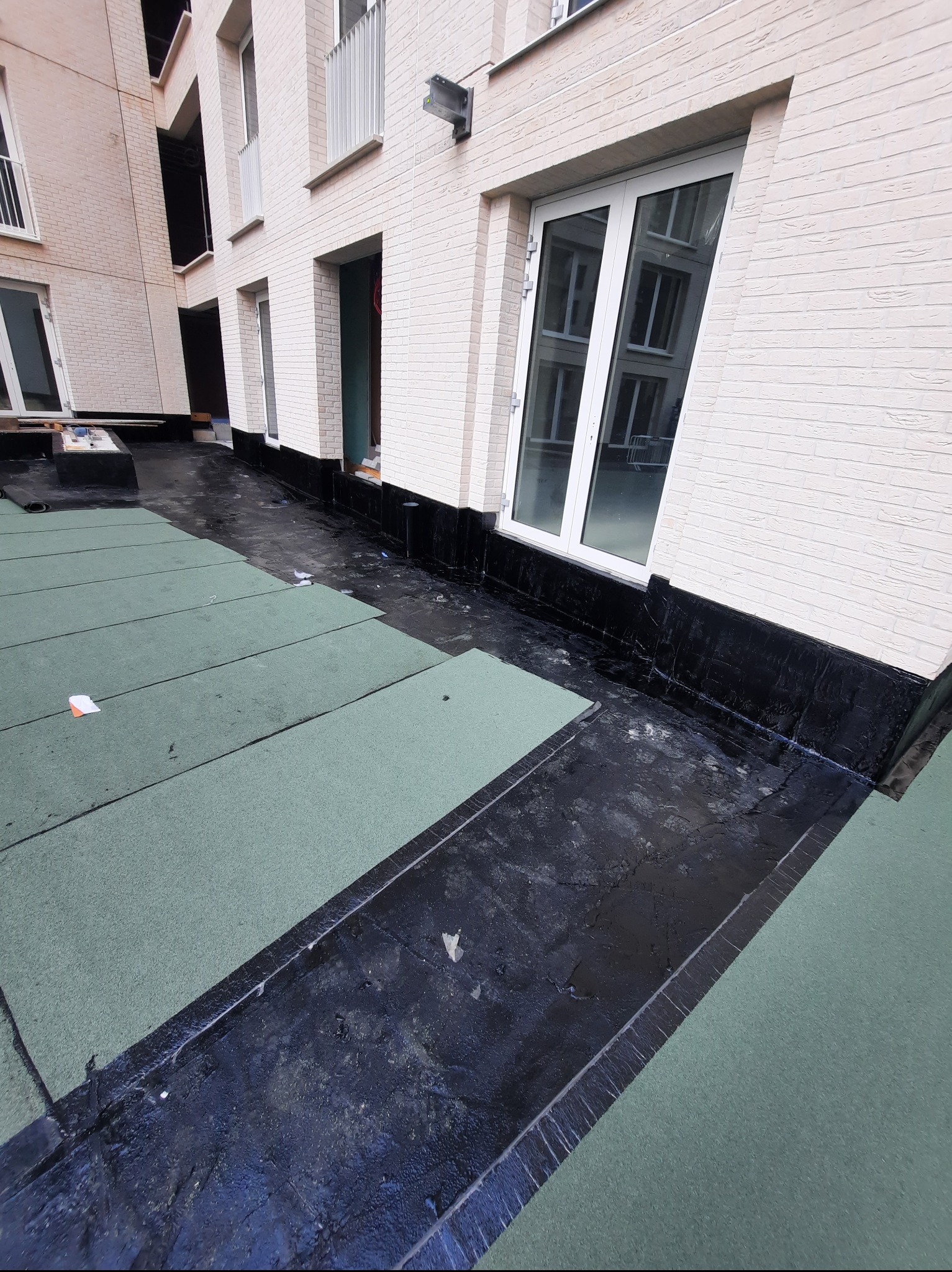 Dach płaski budynku wielorodzinnego z fragmentami zielonej izolacji termicznej i czarną hydroizolacją przy ścianie. Widok z góry ukazuje etap prac wykończeniowych.