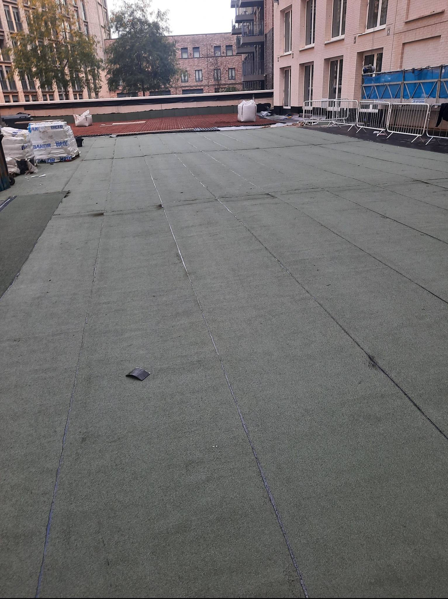 Zielony dach płaski po kryciu papą termozgrzewalną, widoczne zgrzewy. W tle budynki mieszkalne i materiały budowlane, brak osób.