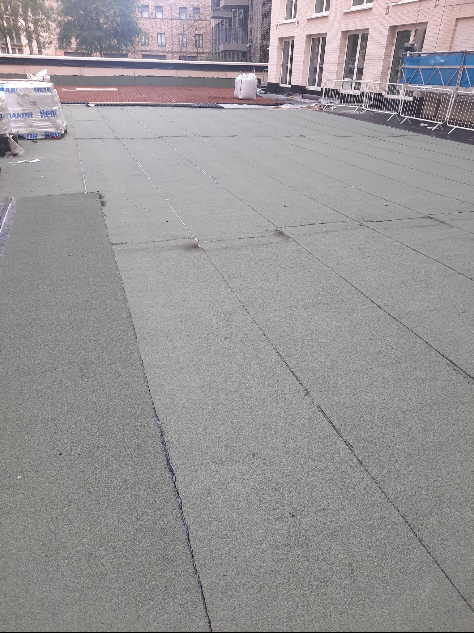 Zielony dach z papy termozgrzewalnej na płaskim dachu budynku wielorodzinnego, widoczne łączenia i fragmenty materiałów budowlanych. Częściowo ukończone krycie.