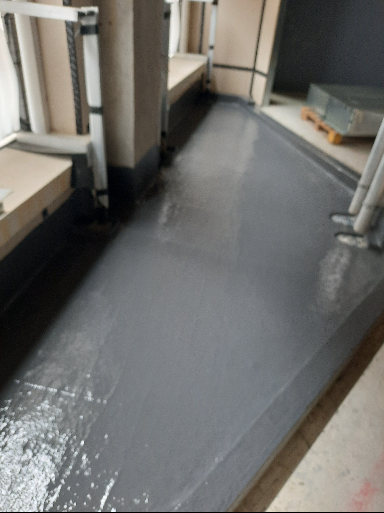 Szara, świeżo wylana posadzka w pomieszczeniu z oknami, widoczne elementy konstrukcyjne i materiały budowlane w tle. Widać mokrą powierzchnię.