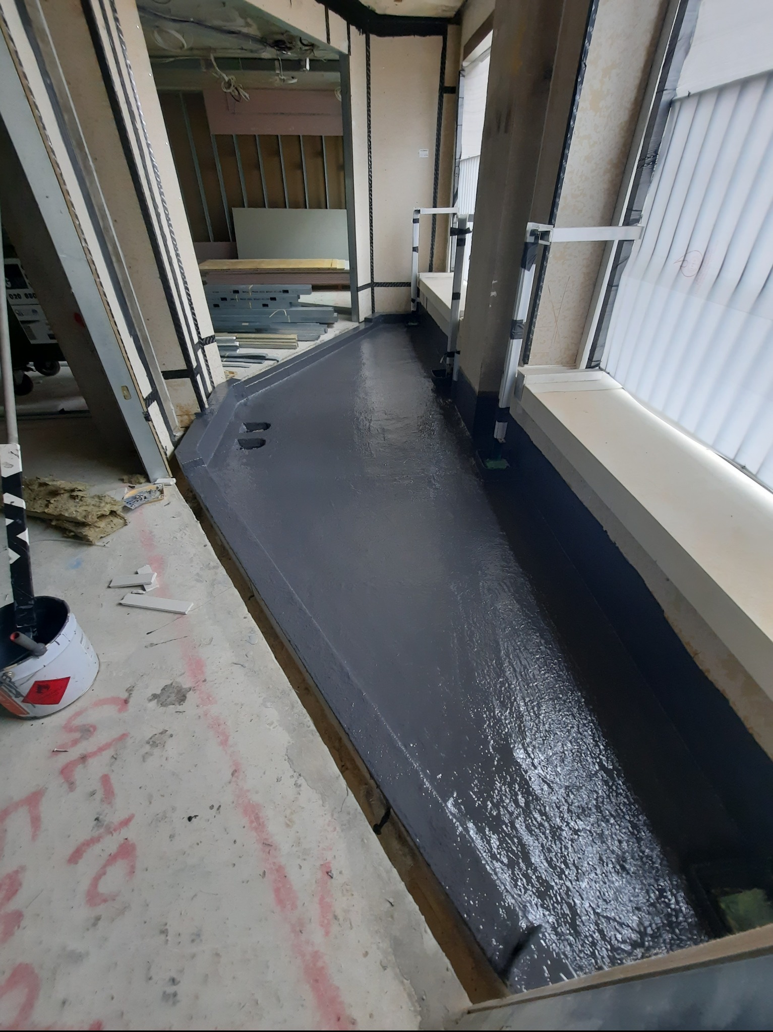 Hydroizolacja balkonu w trakcie prac: świeżo nałożona, ciemnoszara warstwa izolacyjna na podłożu, widoczne okno i ściany w surowym stanie. Wiadro z farbą w rogu.