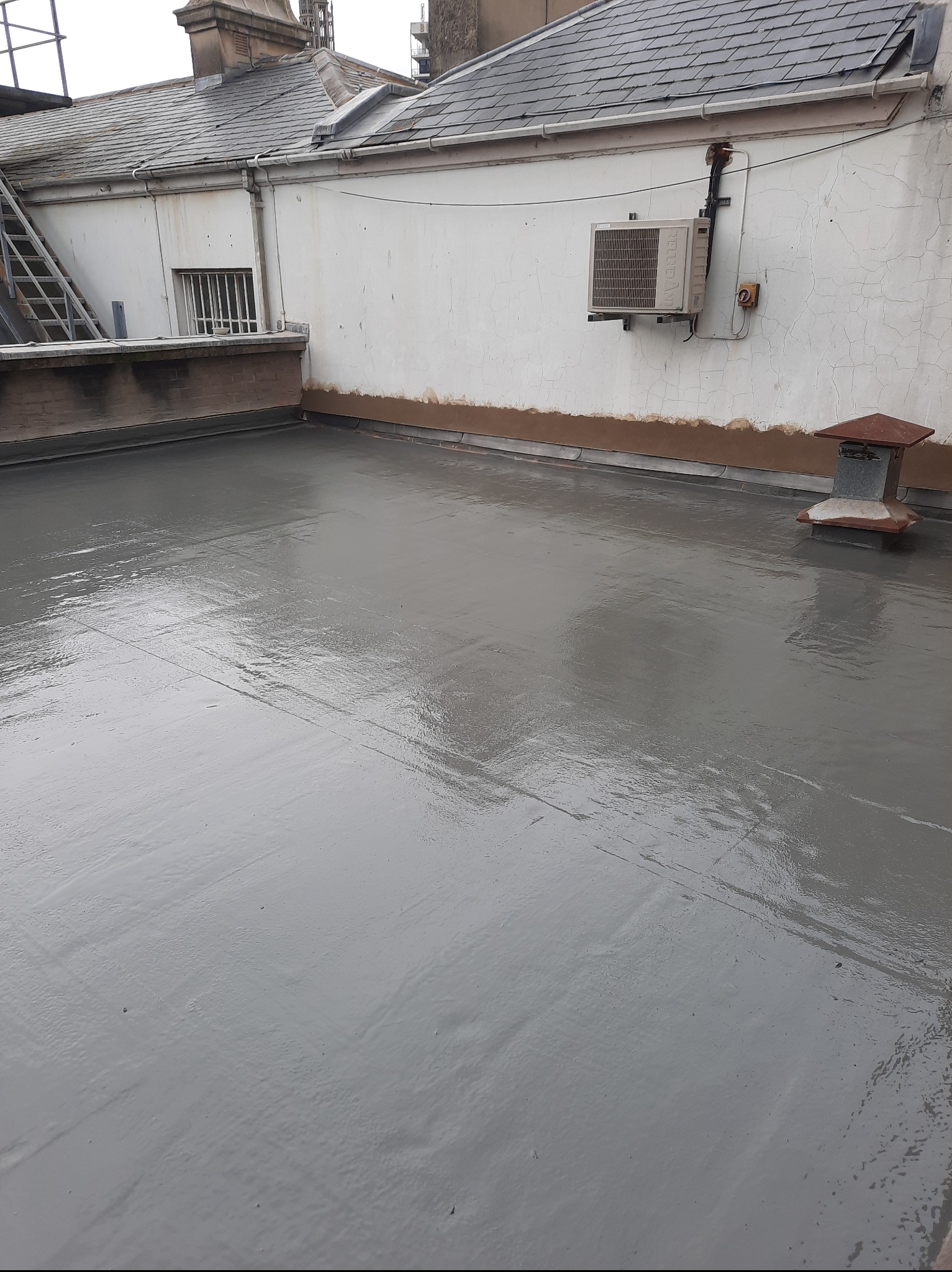 Świeżo położona, szara izolacja na płaskim dachu budynku z kominem. Widoczna ściana budynku z klimatyzatorem i schody przeciwpożarowe w tle.