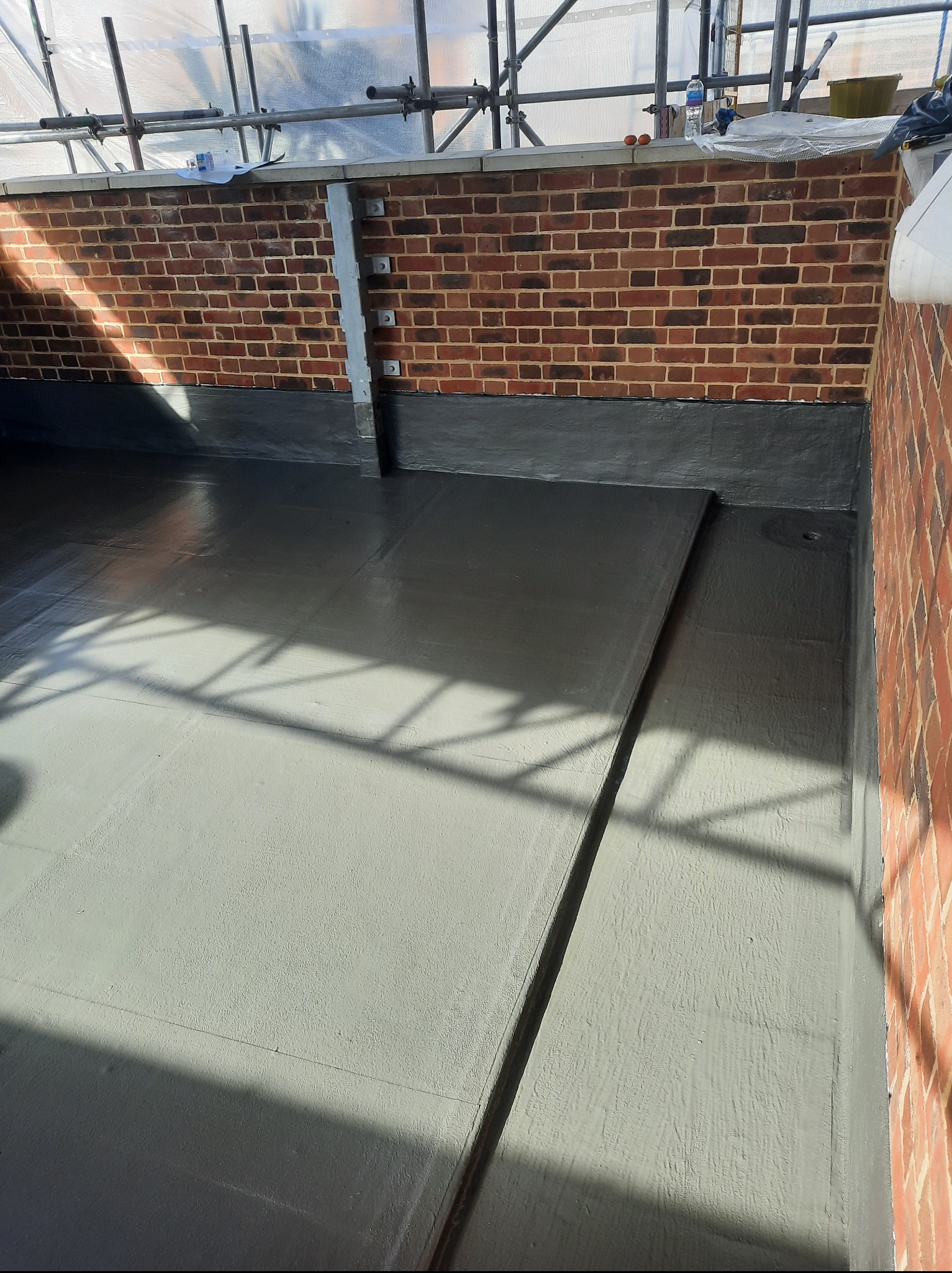 Szare, płaskie pokrycie dachu z ceglaną ścianą i rusztowaniem w tle. Widoczne cieniowanie od konstrukcji rusztowania. Nowe, jednolite wykończenie.