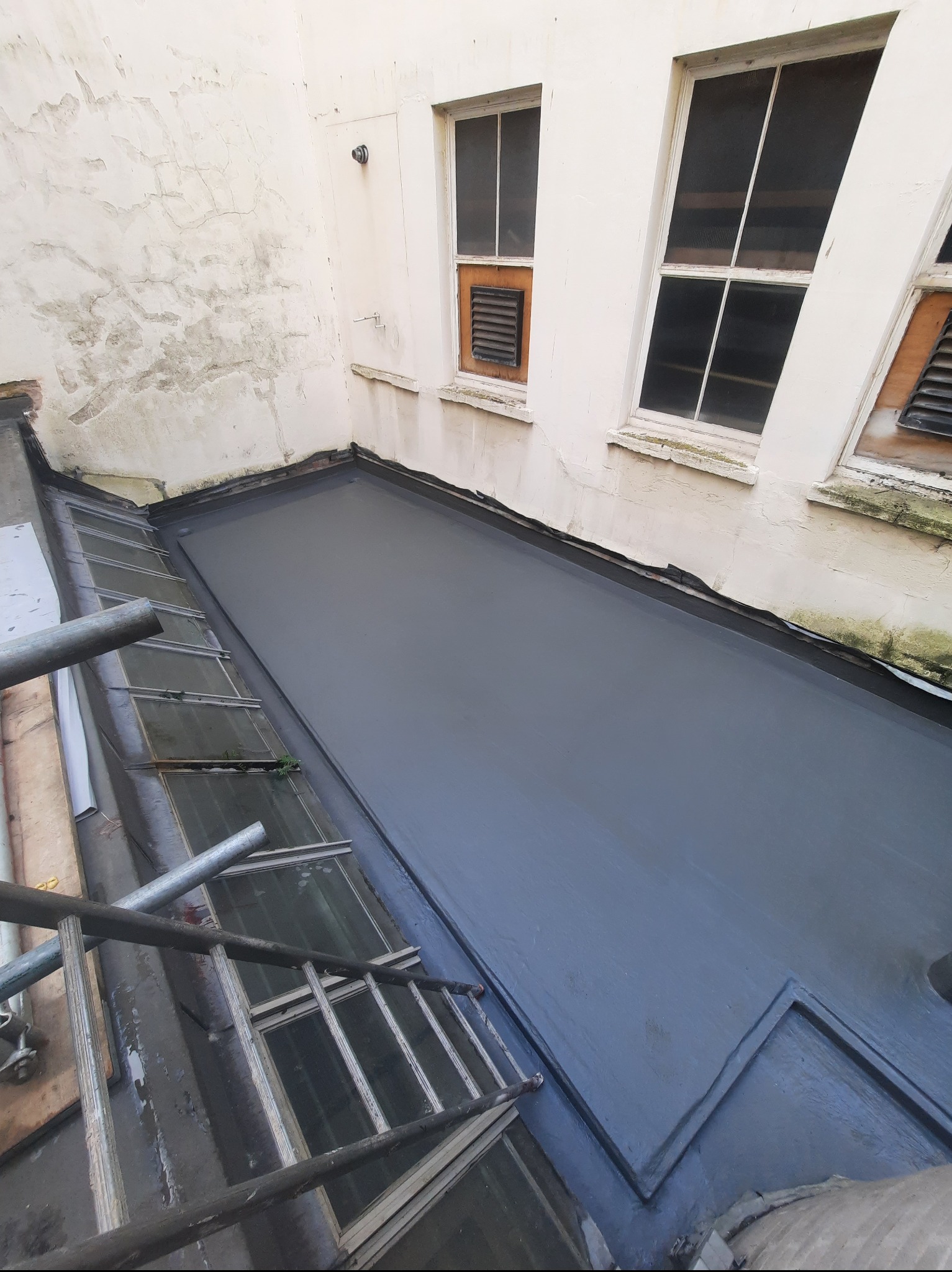 Płaski dach pokryty nową, szarą izolacją bitumiczną, widoczny z góry. Fragment rusztowania po lewej. Biała ściana z oknami w tle.