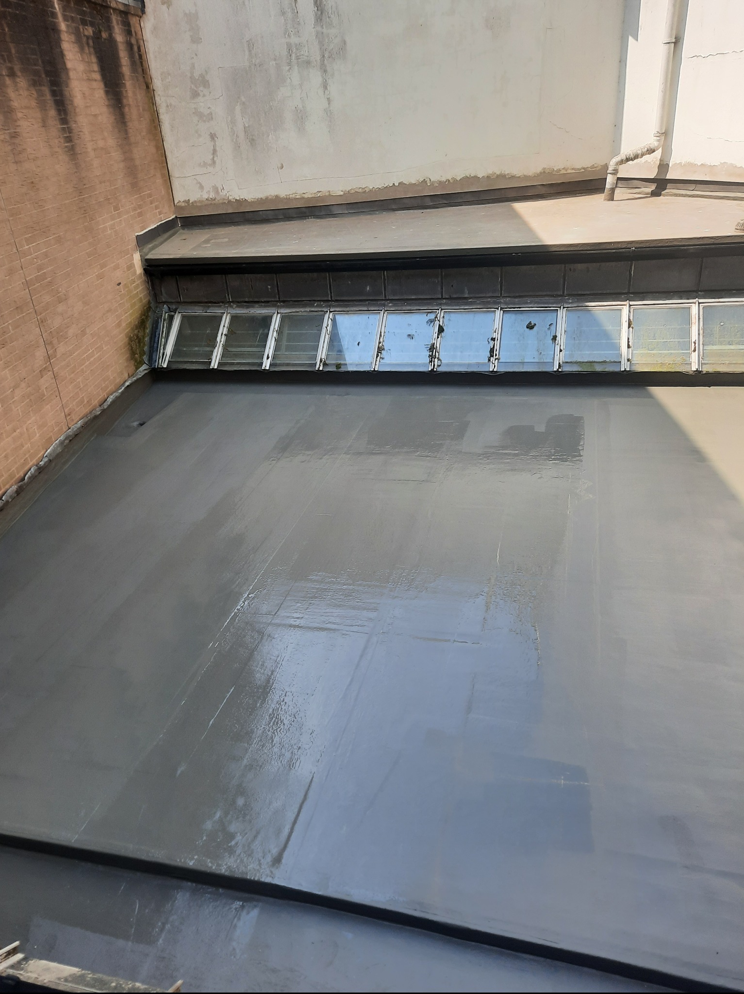 Świeżo pomalowany, płaski dach z oknami dachowymi, widok z góry. Szary kolor farby, ślady pędzla, ceglany mur z lewej strony. Widoczne odbicia światła na mokrej powierzchni.