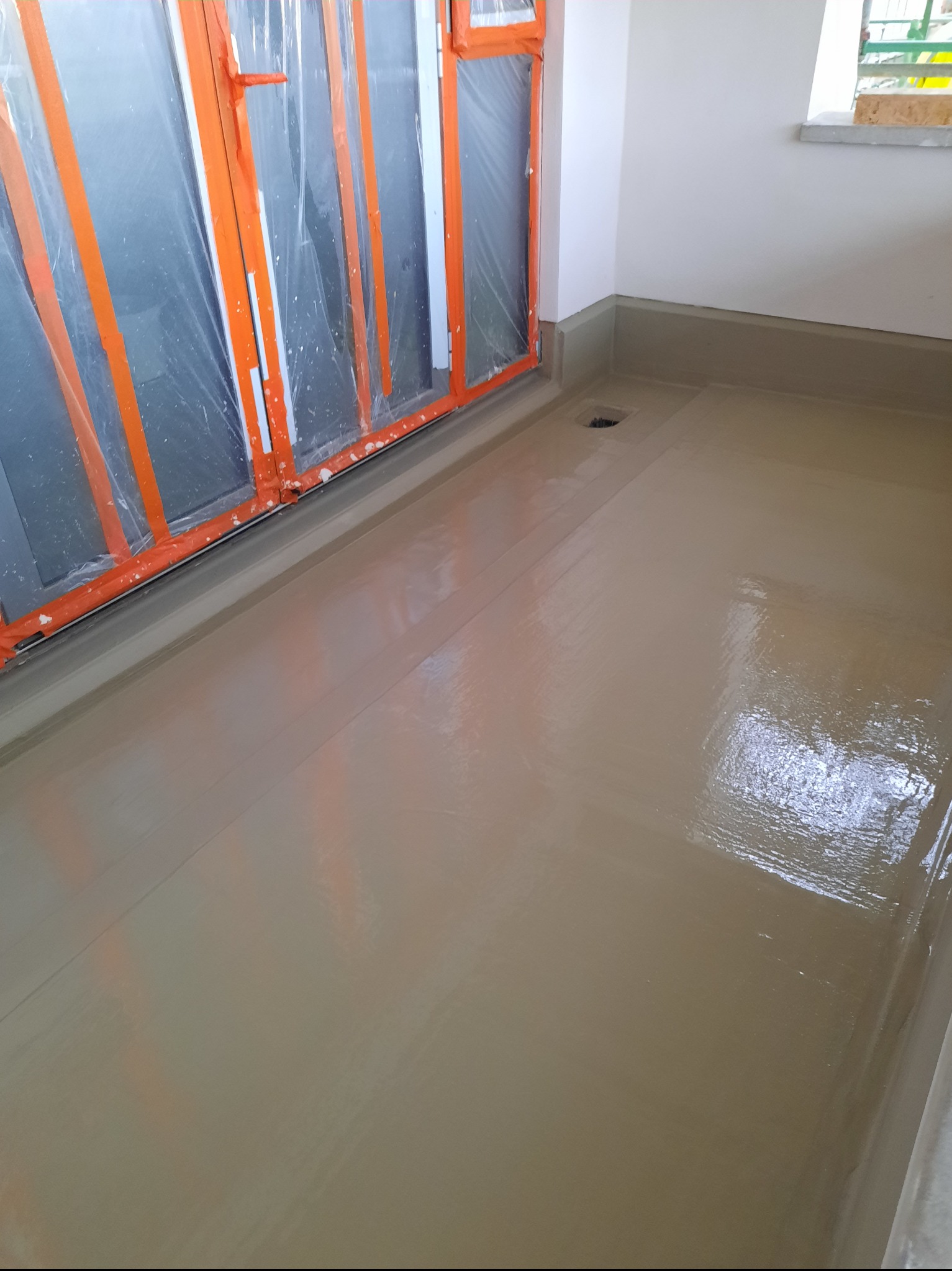 Wnętrze po wylewce samopoziomującej, z zabezpieczonymi folią i taśmą oknami balkonowymi. Widoczny odpływ liniowy i świeżo wylana, gładka powierzchnia.