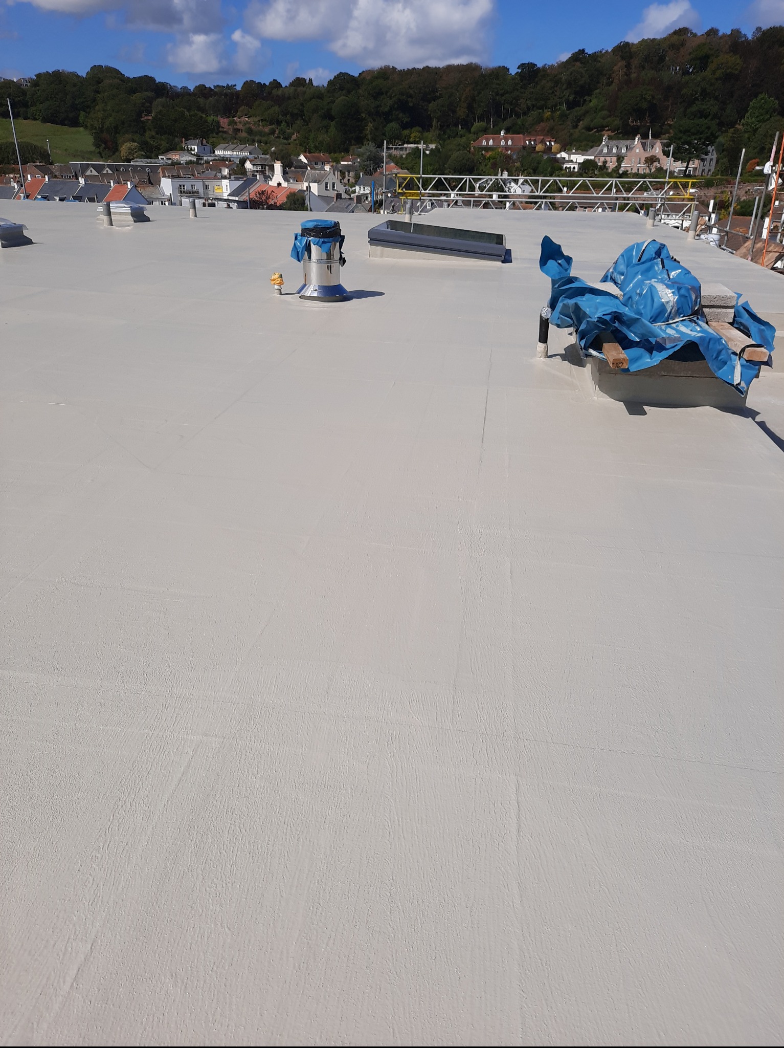 Jasnoszary dach płaski po hydroizolacji, z widocznymi elementami wentylacji i zabezpieczeniami. W tle zabudowa miejska i zieleń wzgórz.