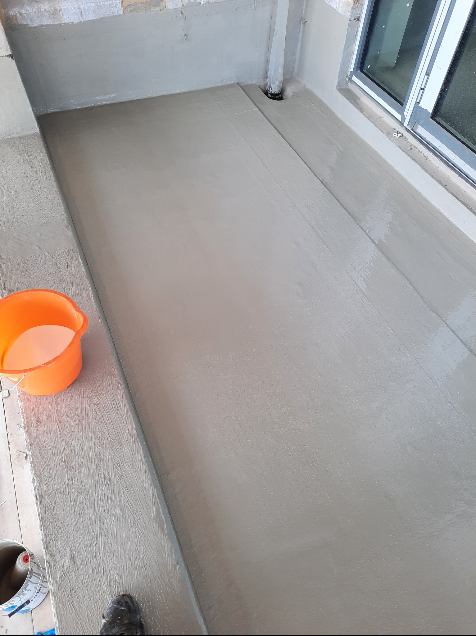 Wyrównana, szara wylewka na balkonie, z widocznym odpływem i oknem. Pomarańczowe wiadro i puszka farby w rogu kadru. Etap prac wykończeniowych.