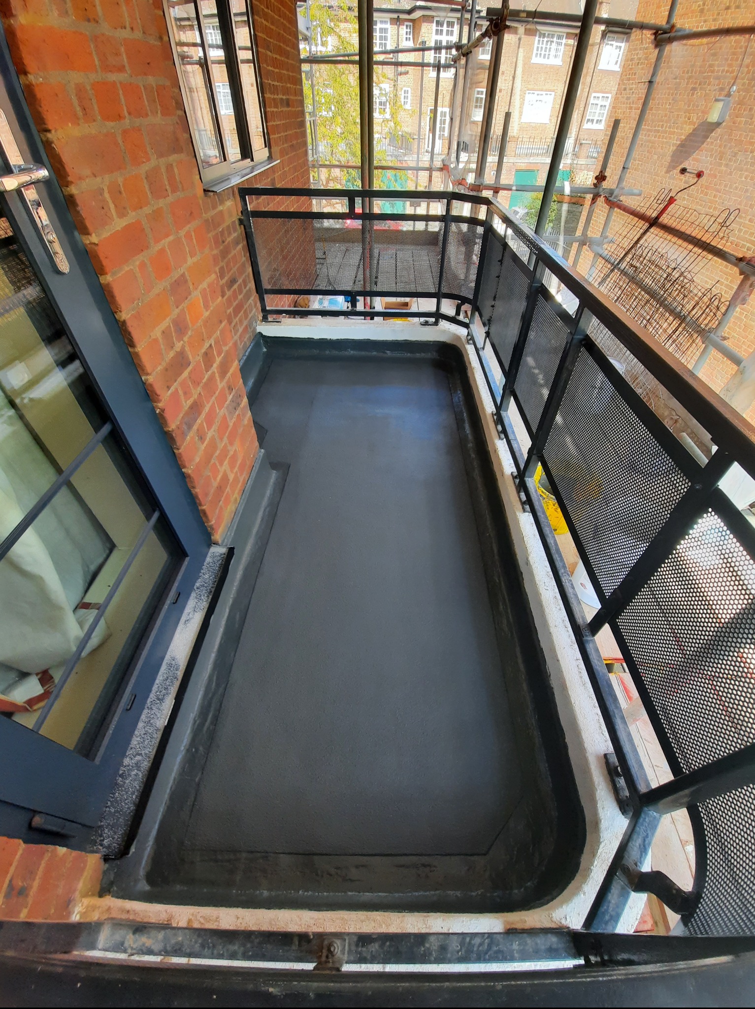 Wykończony balkon z nową, szarą hydroizolacją, czarną metalową balustradą i widokiem na rusztowania i budynki z cegły. Kąt z góry.