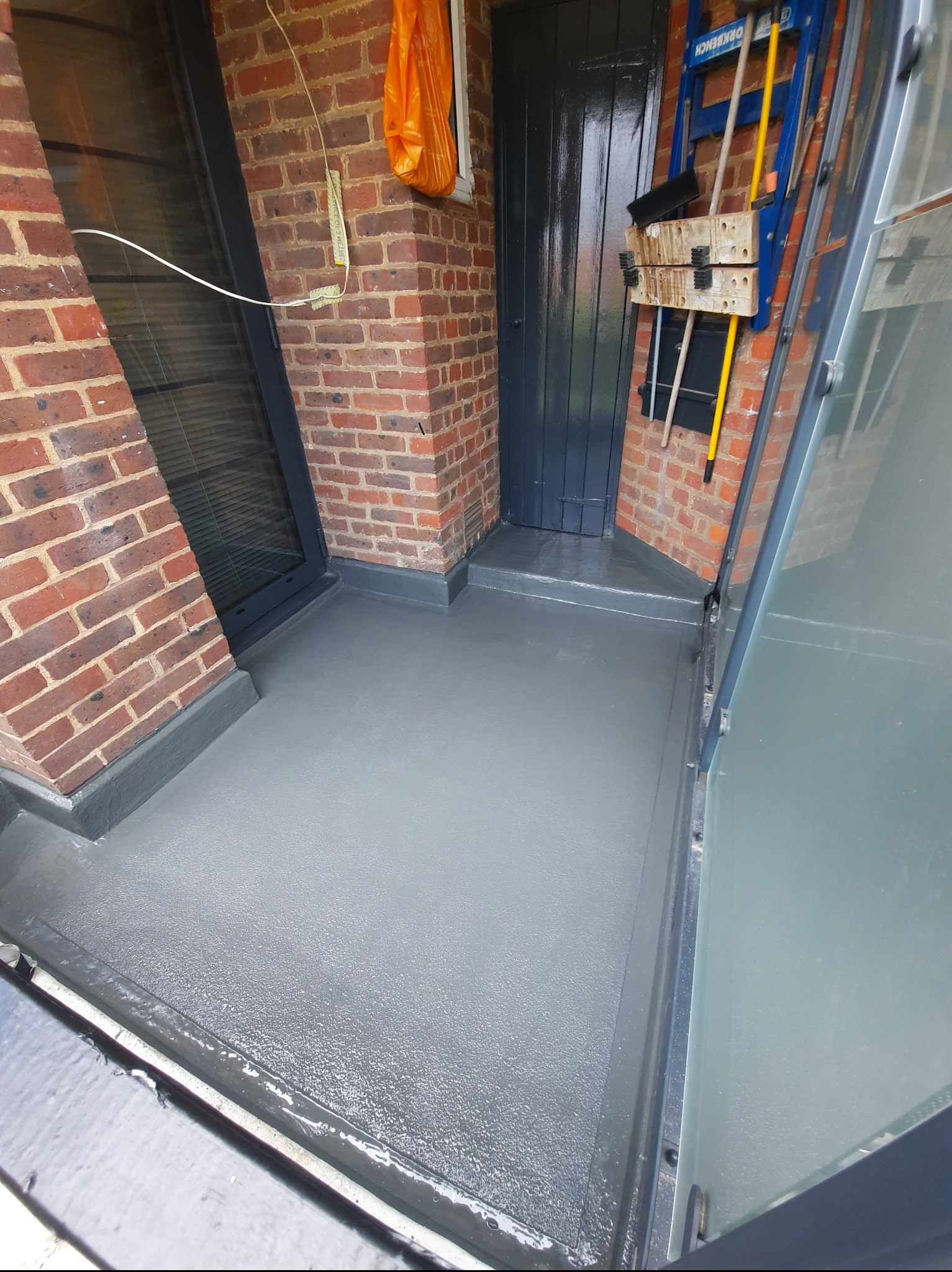 Szara, świeżo pomalowana płaska powierzchnia dachu przylegająca do ceglanej ściany z oknem i drzwiami. Widoczne narzędzia i drabina.