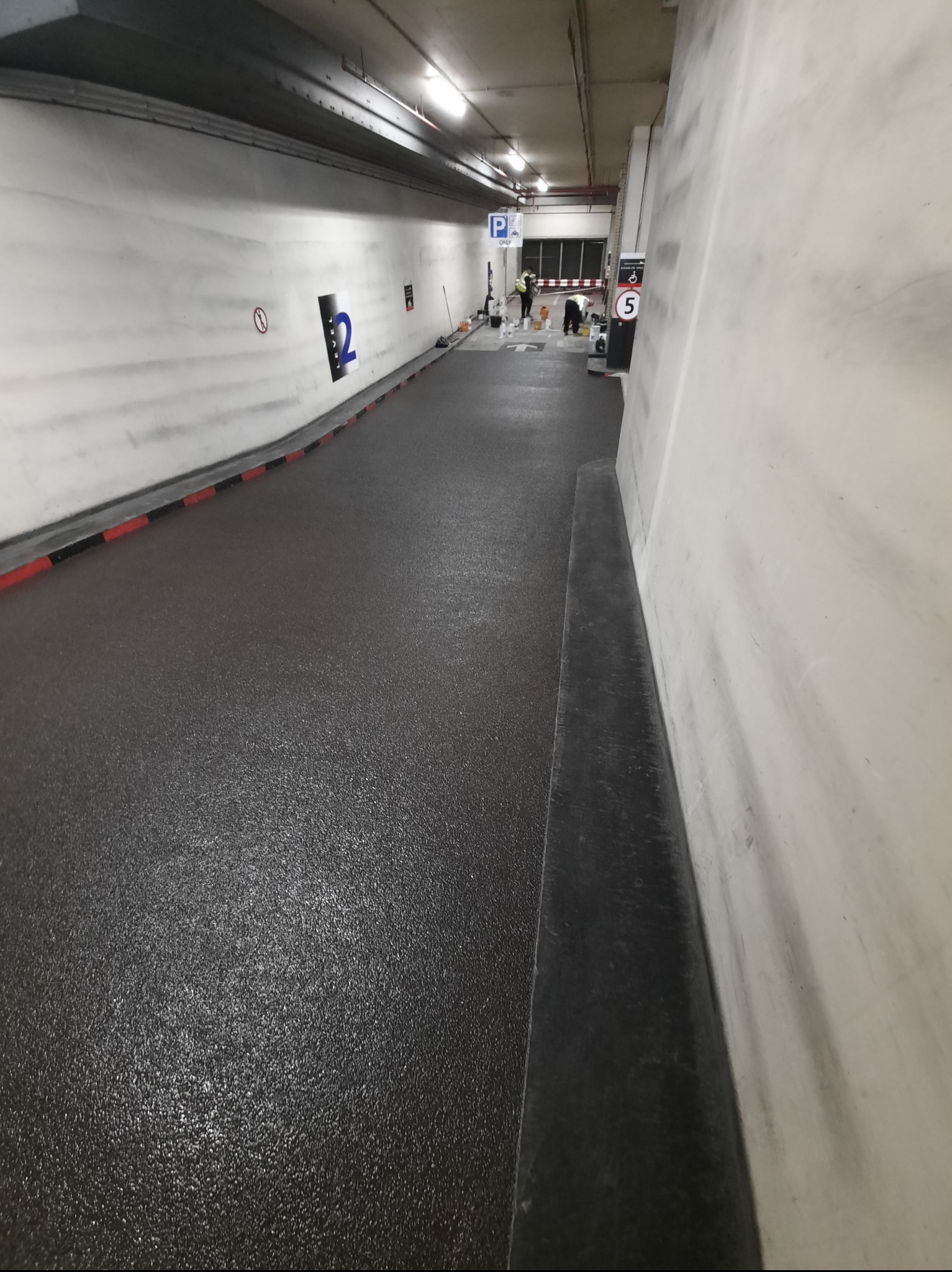 Prace wykończeniowe w garażu podziemnym: świeżo pomalowana posadzka w ciemnym kolorze, widoczne oznakowania poziomu i stanowiska, ekipa w trakcie prac malarskich.